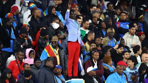 Reuters Michel Nkuka Mboladinga dressed as Patrice Lumumba in a blue suit jacket and red trousers with his hand raised in the middle of Congolese fans in a stadium in Rabat.