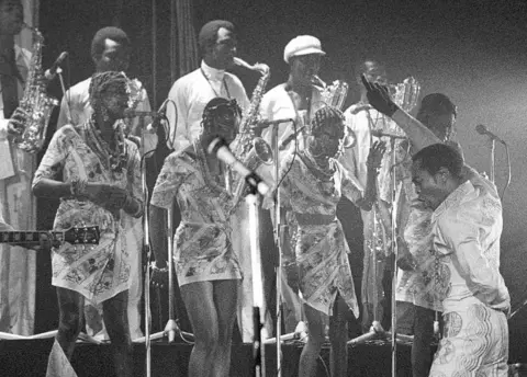 Redferns/Getty Images Black and white shot of Fela Kuti with his back to the audience and facing his back-up singers and band. He is singing with one hand up and finger pointing and the other hand behind his back in a pose like a torero.