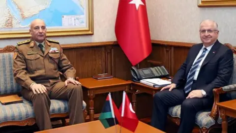 Reuters Libya's Gen Mohammed Ali Ahmed al-Haddad, wearing a brown military uniform, meets Turkish Defence Minister Yasar Guler in Ankara, wearing a black suit. Both are seated on sofas in a room with a map and flags of their countries. Photo: 23 December 2025