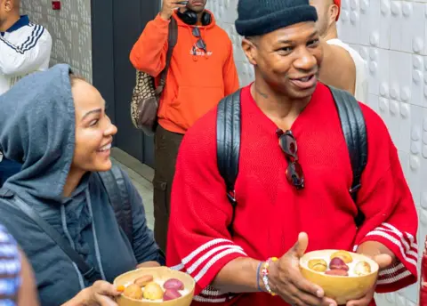 Mohamed Finando / Oxygen Africa US actors Meagan Good and Jonathan Majors being welcomed at Conakry Gbessia International Airport.  They were presented with kola nuts. Kola nuts hold deep cultural significance in West Africa, symbolising hospitality, peace, friendship, and life.  They are used in crucial social rituals like weddings, funerals, and negotiations.   The couple are smiling, chatting and wearing relaxed clothing.
