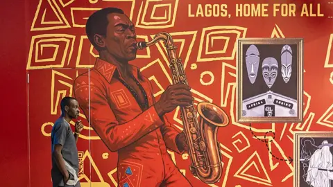 AFP/Getty Images A man walks in front of mural in Lagos of Fela Kuti in a red jumpsuit playing a saxophone, with the words 'Lagos, Home For All'.
