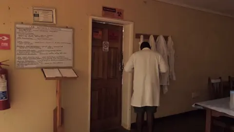 TRACE, Simon Dures A man in a white coat stands to the right of the picture with his back to the camera. He is in a dark corner of a room with walls painted in a light yellow colour. The room has a noticeboard on the wall to his left beyond a door. At the top of the noticeboard a sign reads "Lab actions and Projects".