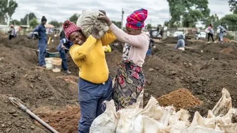 AFP/Getty Images A woman grimaces as she is helped by another woman to load a sack of soil on to her shoulder in a patch of land where artisanal miners look for gold outside Springs.
