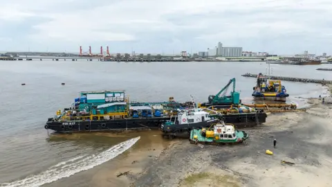 AFP via Getty Images An aerial view of the city of Toamasina, on the east coast of Madagascar.  Three boats are grounded on a sandy shoreline beside a wide bay. Machinery and equipment sit on the vessels, with one tugboat tilted at an angle on the beach. A person walks nearby, and industrial buildings and cranes are visible across the water in the distance.