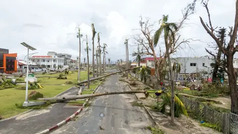 AFP via Getty Images An aerial view of the city of Toamasina, on the east coast of Madagascar.  A road lined with palm trees heavily damaged by Cyclone Gezani, with several trees snapped or uprooted and lying across the street. Debris and fallen branches cover the area, surrounded by buildings, under a cloudy sky.