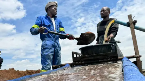 Thuthuka Zondi/BBC A man in blue overalls holds a spade as he washes gold granules with a colleague wearing a black jumper.