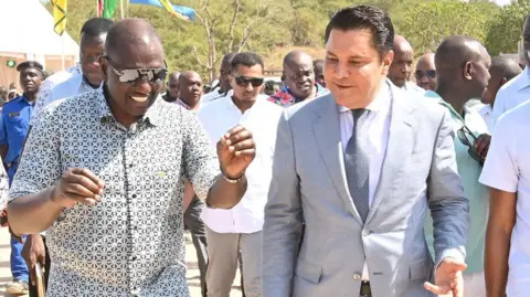 State House Kenya President William Ruto, wearing a checked shirt and sunglasses, walks and talks with businessman Rostam Aziz, who is wearing a grey suit