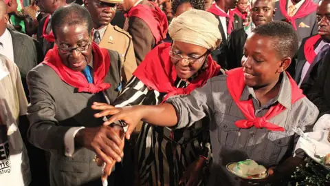 Gallo Images/Getty Images Robert, Grace and Bellarmine Mugabe are standing together, smiling and wearing red neck scarves as they jointly cut a cake.