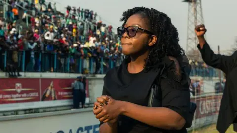 AFP via Getty Images Bona Mugabe is wearing sunglasses and dressed in black has her hands clasped in front of her. A crowd of people in a stadium stand can be seen out of focus behind her.
