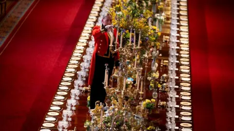 PA Media A member of Royal Household staff lights a candle during table preparations in St George's Hall, they wear a red jacket and black trousers. the long table is decorated in lavish floral arrangements and candle sticks. A row of plates and  cuttlery line the edges of the table.