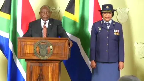 Reuters Cyril Ramaphosa stands at a presidential podium flanked by two South African flags. Next to him, in a police uniform, is Lt-Gen Puleng Dimpane.