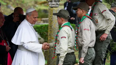 Reuters Pope standing on the left, shaking hands with a male scout. There are two others next to him. They are standing outdoors. Behind the pope is a man holding a camera up to his face.