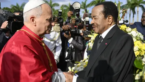 EPA/Shutterstock Pope Leo in a red cape shakes the hand of President Paul Biya in a black suit with a white dotted tie. Behind them are banks of photographers and a floral arrangement.