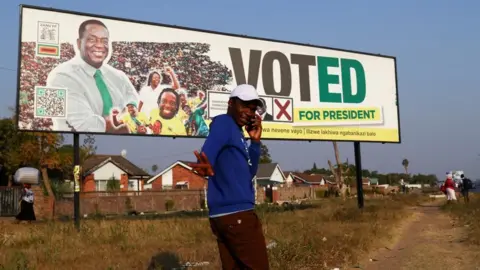Reuters A man gestures while speaking on a cellphone as he walks past a banner of the ruling ZANU-PF party's President Emmerson Mnangagwa ahead of the presidential elections in Harare, Zimbabwe, August 20, 2023