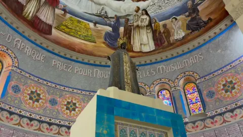 A statue of a black Madonna shot from below. Behind it is an inscription in French on a tiled wall saying, in French, 'Our Lady of Africa pray for us and the Muslims'