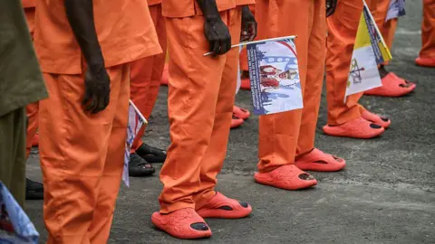 AFP via Getty Images Prisoners wearing orange uniforms holding flags with the Pope's face. You can only see their legs and orange sandals.