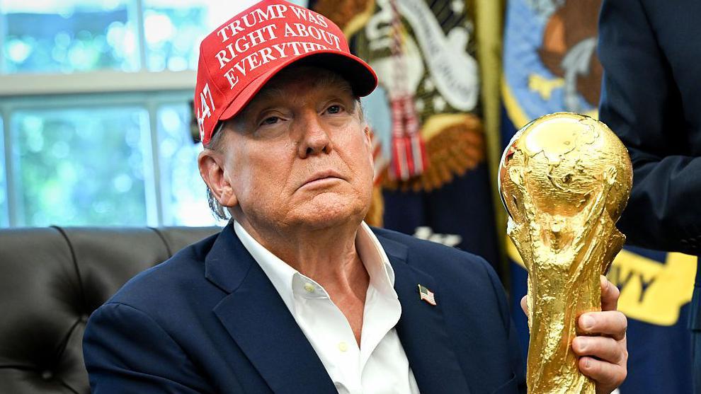 US President Donald Trump visible from chest up holds the Fifa World Cup trophy while sitting in a black chair in the Oval Office in the White House. He wears a blue suit jacket bearing an American flag lapel badge, an open-necked white shirt and a red baseball cap bearing the message 'Trump was right about everything'. Out of focus in the background are a window and two flags