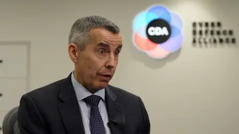 The head and shoulders of a man with short grey hair, wearing a blue jacket, shirt and tie, with a CDA logo blurred in the background.