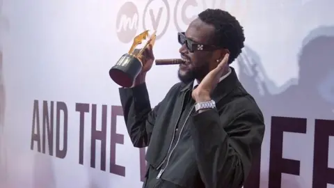 BBC/ MOBOLAJI OLATUNDE  Bucci Franklin, in a black outfit and wearing shades, is on the stage with his award. 