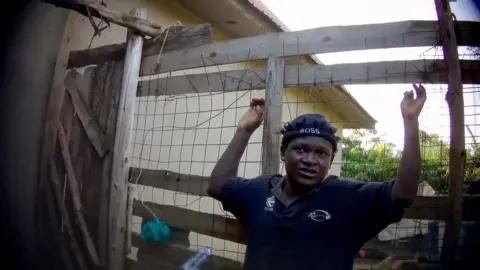 A frame from undercover filming showing Charles Lubajja in a blue Boss-brand cap and T-shirt leaning against a wire fence. He is holding his arms up in a cactus shape.   