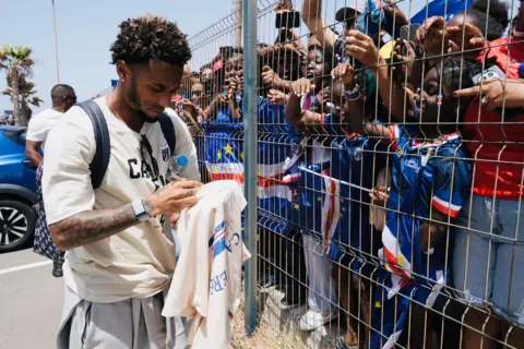 Michel Mvondo/BBC Captain Ryan Mendes signs a white shirt, while separated from fans by a metal fence