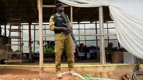 Reuters An armed officer in khaki uniform and black stab vest stands guard in front of a makeshift room.