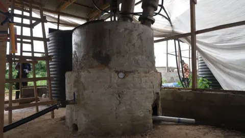 A concrete cylinder with a temperature gauge on the side sits in the middle of a room - pipes are coming out of the top and a grey tube is coming out of the bottom. Plastic sheeting can be seen in the place of a wall to the right.  