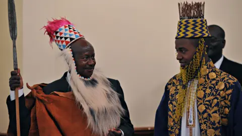 AFP via Getty Images Ugandan President Yoweri Museveni (L) wearing traditional Tooro kingdom ceremonial head gear smiles at Ugandan King Oyo in 2010