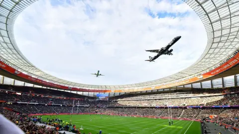 Gallo Images/Getty Images A fish-eye view from the stands of a rugby stadium looking up at the sky as two planes fly in close formation above.