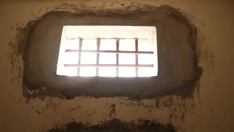 A small window with bars across it lets through light. It is shot from below.