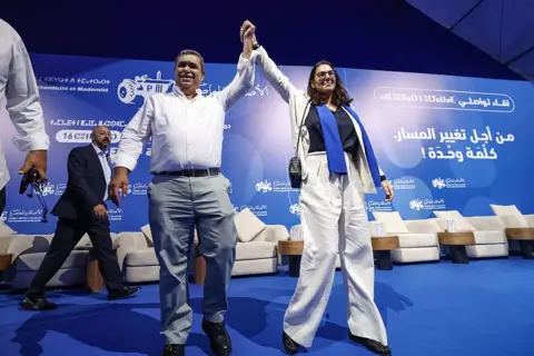 AFP via Getty Images El Mansouri holding hands with a Pam colleague during one of the part's rallies. She is wearing a white suit and standing on a blue platform featuring Pam's logo in several languages.