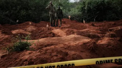 AFP via Getty Images Two men in military uniform stand on red earth where several large holes have been dug up. Behind them tress can be seen. In the foreground is a yellow banner which reads: Crime scene do not cross