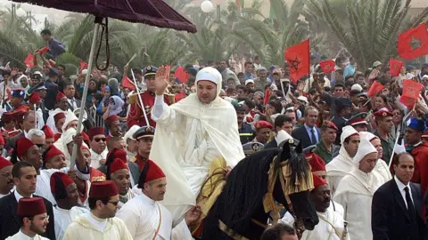 AFP via Getty Images Morocco's King Mohammed VI, wearing a long white traditional robe, rides a horse amid a large crowd of security personnel, military officers, officials and civilians, waving to the crowd.