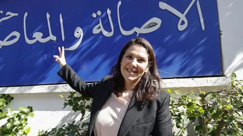 AFP via Getty Images El Mansouri, wearing a black blazer over a white shirt, stands in front of her party's slogan, written in Arabic in white lettering against a blue background.