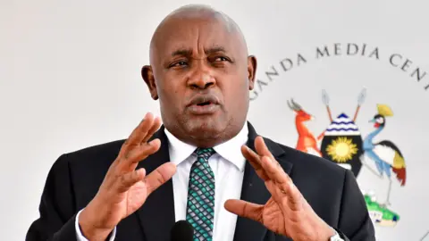 Reuters Uganda's Health Minister Chris Baryomunsi, in black suit and white shirt, gestures as he speaks. There is a black microphone in front of him and a banner with words Uganda Media Centre and a Ugandan coat of arms behind him.