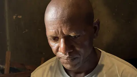 Mohamed Madi / BBC A close-up shot of a bald man wearing a white T-shirt looking down
